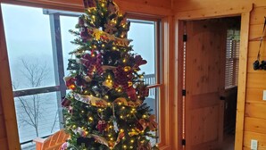Un árbol de Navidad decorado con luces y adornos en una habitación con paredes de madera y un ventanal grande.