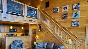 Interior acogedor de una cabaña con escalera de madera, espacio de cocina, zona de estar con sofá y butaca estampada, y diversas decoraciones en las paredes.