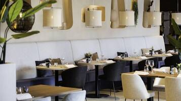 A modern restaurant with round tables and chairs, a white cushioned bench seating area, and wall-mounted pendant lighting.