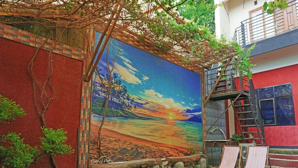 Un mural de una escena de playa en una pared de edificio, con un banco de madera y dos sillones de descanso al frente.