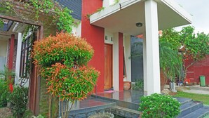 Una casa moderna con fachada roja, un porche cubierto y un pequeño jardín con diversas plantas y una fuente de agua.