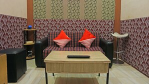 Una sala de estar con un sofá, dos almohadones rojos, una mesa de centro de madera, un control remoto, un altavoz negro y un papel tapiz estampado.