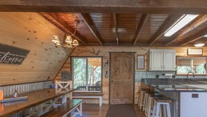 Interior rústico de una cabaña con una zona de estar con banco de madera, una cocina con armarios blancos y una puerta grande de madera.
