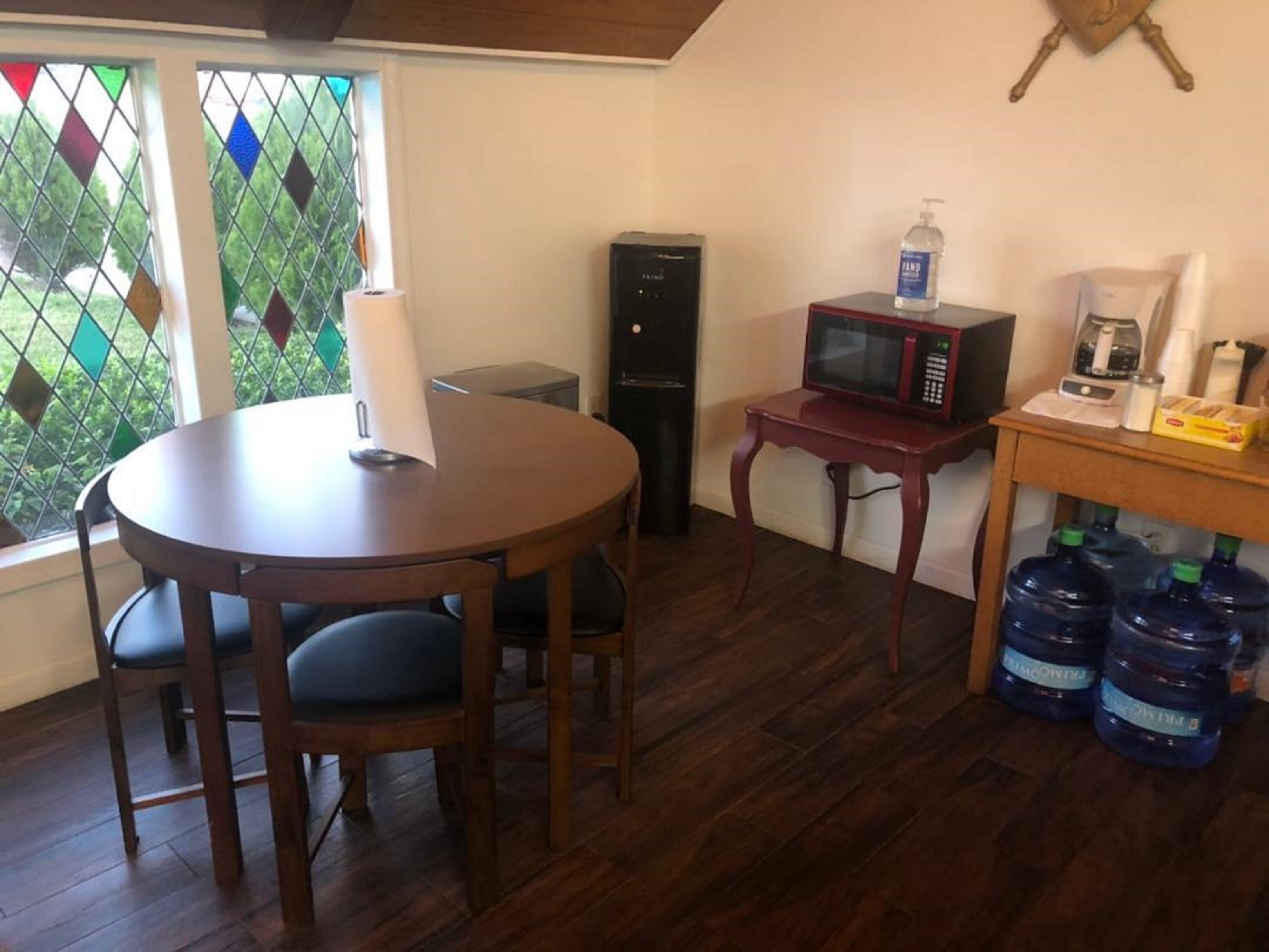 A room with a round wooden table, two chairs, a microwave, a small refrigerator, and a window with stained glass.