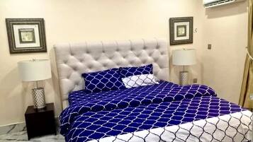 Une chambre à coucher avec une tête de lit capitonnée, un lit recouvert d’une couvre-lit à motifs bleus et blancs, deux lampes de chevet et des tableaux encadrés accrochés aux murs.