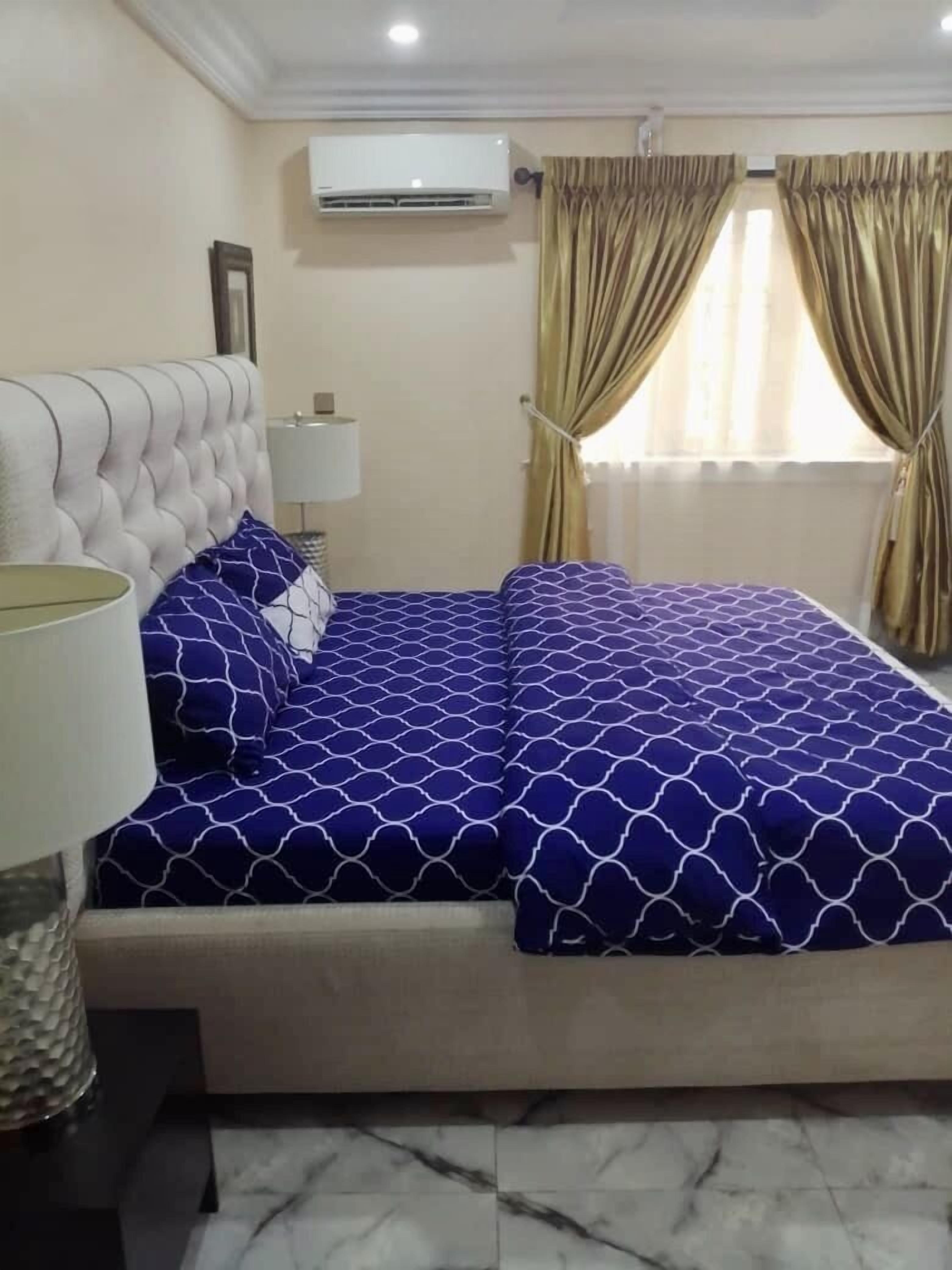 A bedroom with a tufted headboard, a bed with a blue patterned bedspread, a bedside lamp, an air conditioning unit, and a window with curtains.