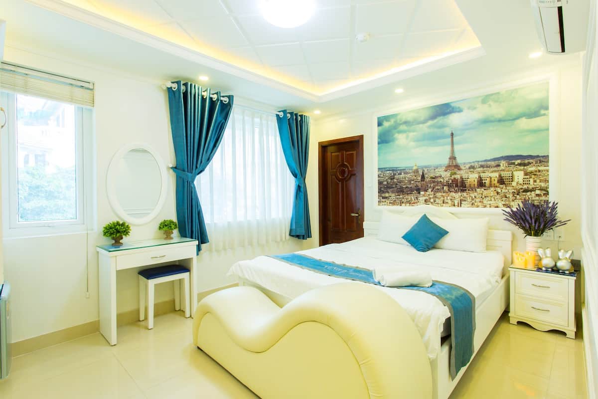 Un dormitorio con cama, escritorio, silla, espejo, mesita de noche con velas y un cuadro de un paisaje urbano en la pared.