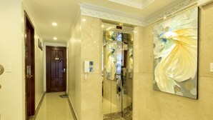 A hallway with a marble floor, a wooden door, and a glass elevator. - Venice Hotel (Ho Chi Minh City)