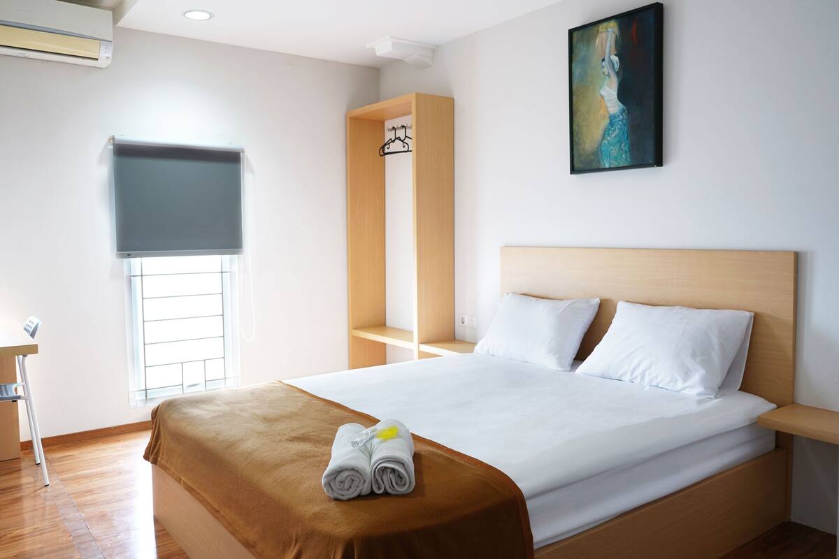 A hotel room with a bed, a desk, a window with blinds, a painting on the wall, and a wooden headboard.
