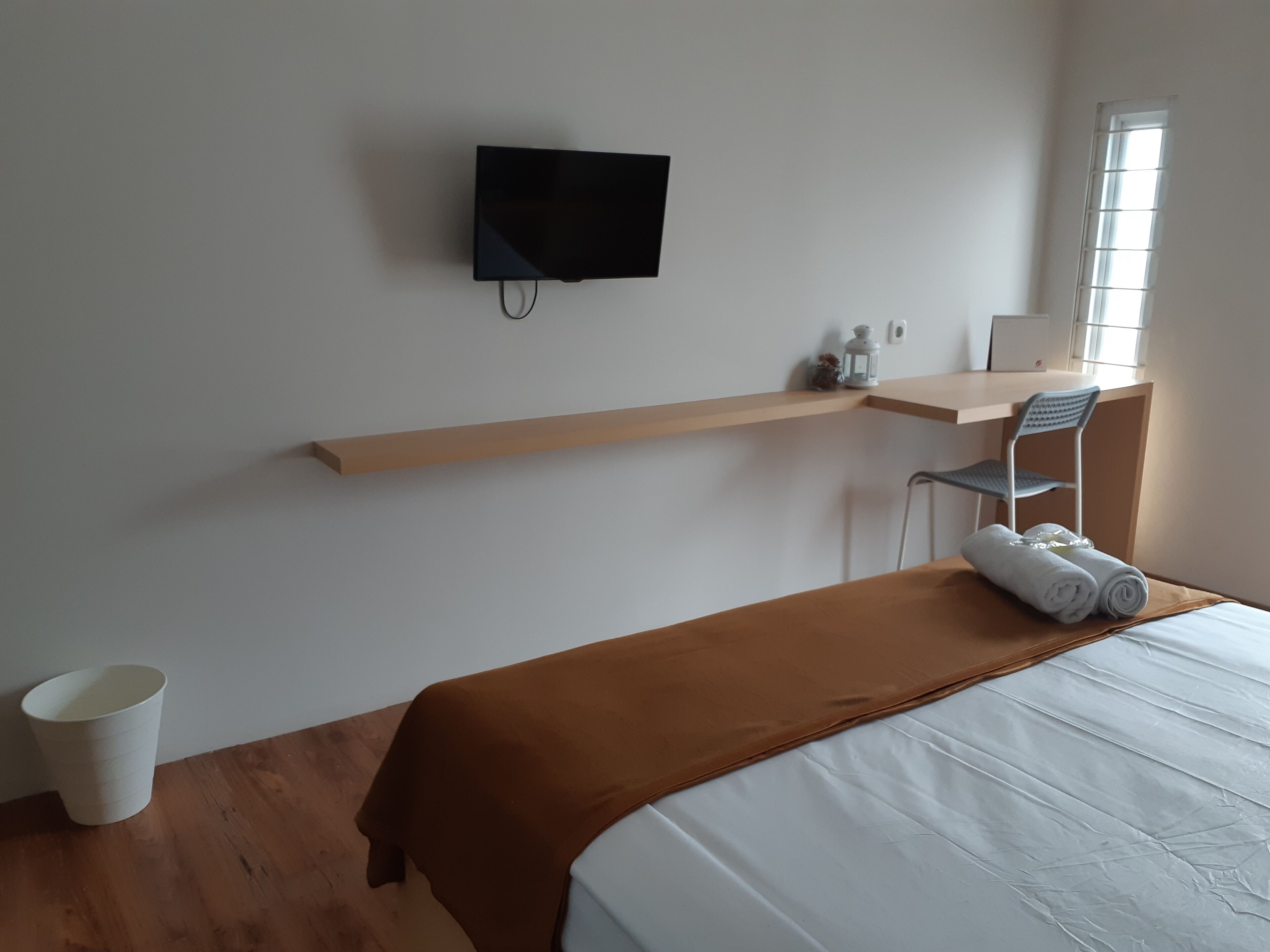 A hotel room with a bed, a television mounted on the wall, a wooden desk, a chair, and a window.