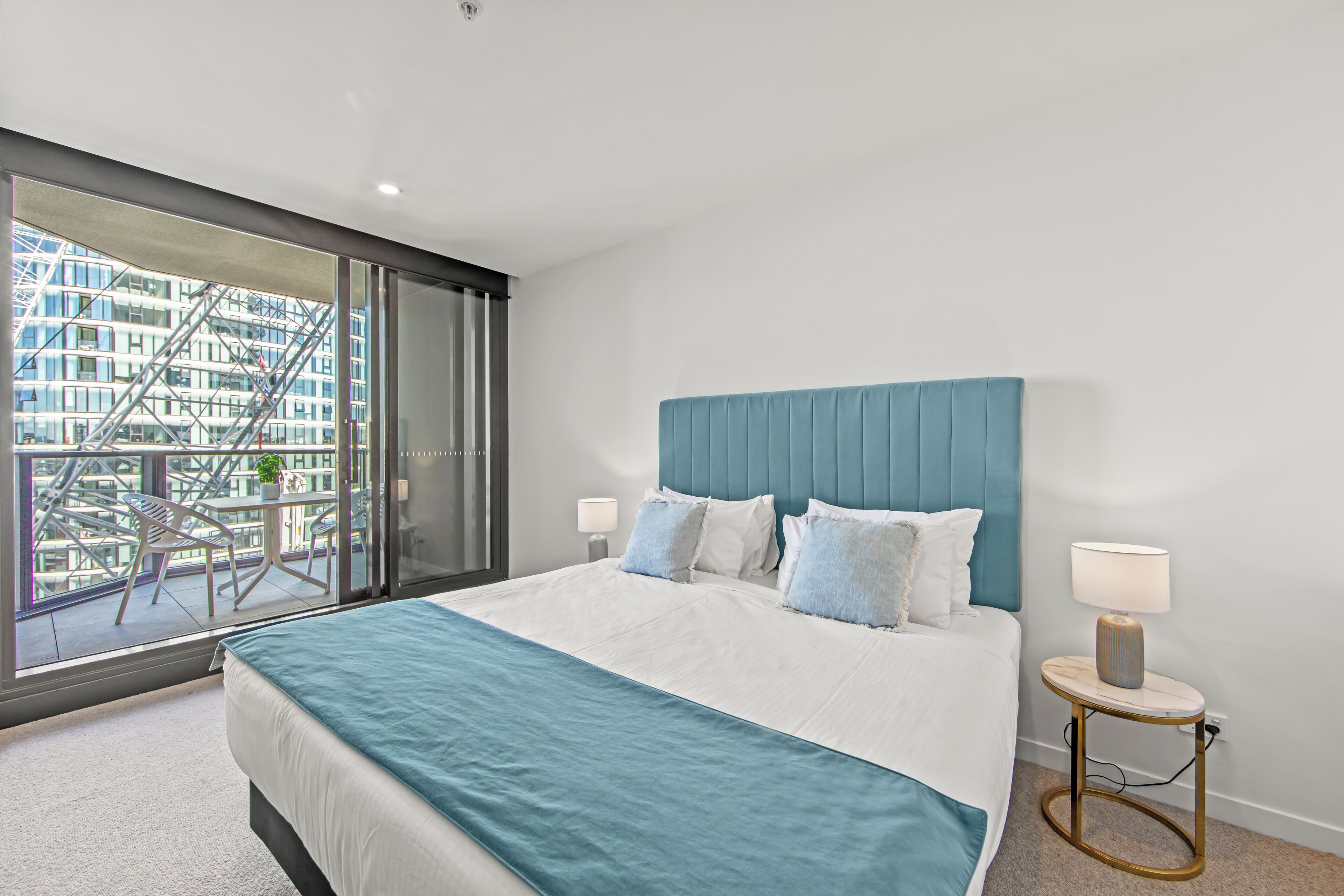 Una habitación de hotel moderna con una cama grande, vistas de la ciudad a través de ventanas de piso a techo, y un balcón con mobiliario exterior.