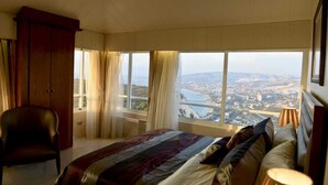 A bedroom with a large bed, a chair, a wardrobe, and a view of a cityscape through large windows. - Theodos Lodge Hotel By Hansa (Batroun)