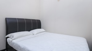 Una cama pequeña y bien hecha con ropa de cama blanca en una habitación minimalista con paredes blancas y una única lámpara de pared.