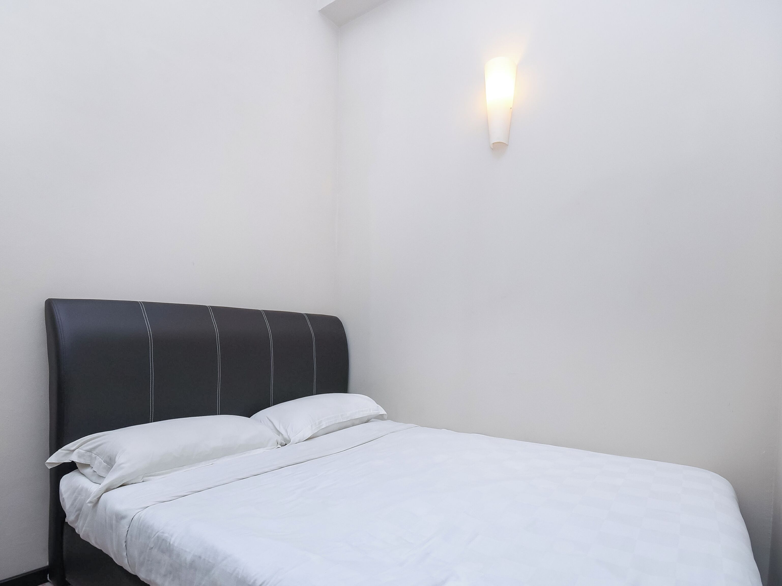 A single bed with a dark headboard and white bedding in a room with a plain white wall and a wall-mounted light fixture.