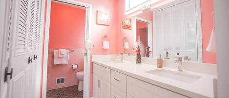 Un baño con pared rosa, muebles blancos, un espejo grande y un inodoro.