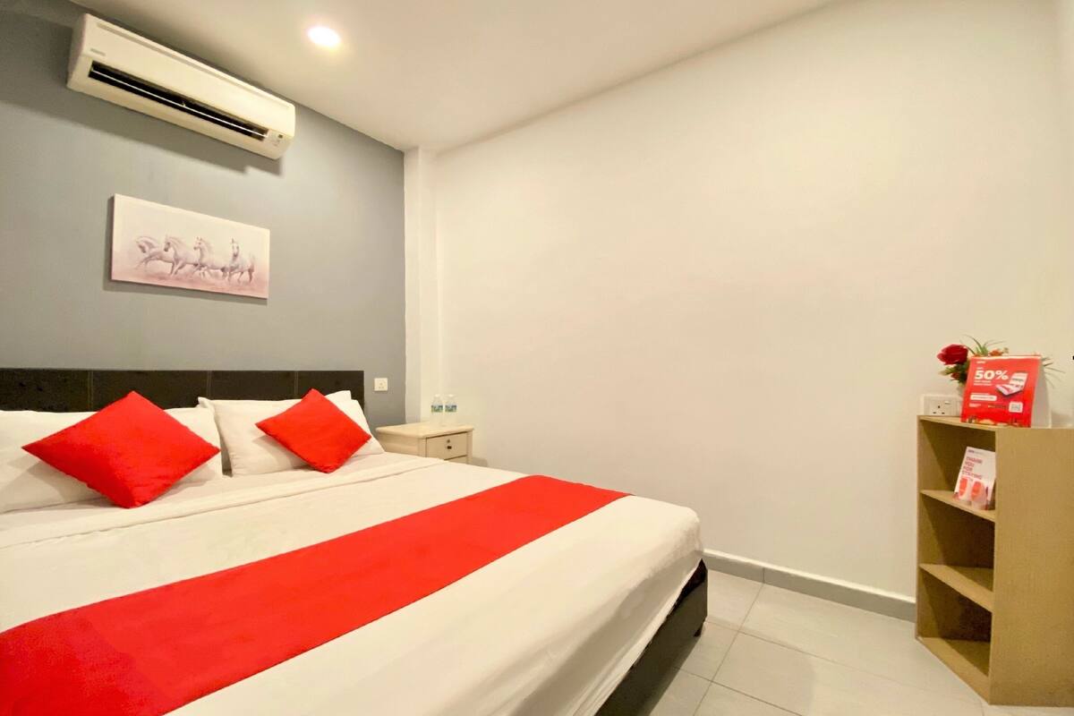 A hotel room with a bed, red pillows, a white bedspread, an air conditioner, a nightstand, a small table, a shelf with a red box, and a painting on the wall.