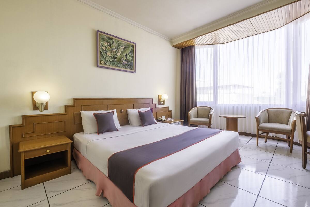 A hotel room with a large bed, bedside tables, a nightstand, a chair, a small table, and a window with curtains.