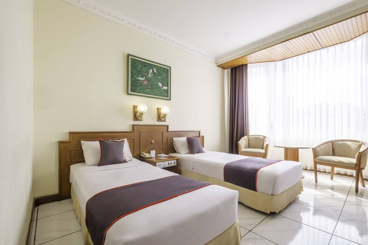 A hotel room with two beds, a wooden headboard, a painting on the wall, and a view of the city through large windows.