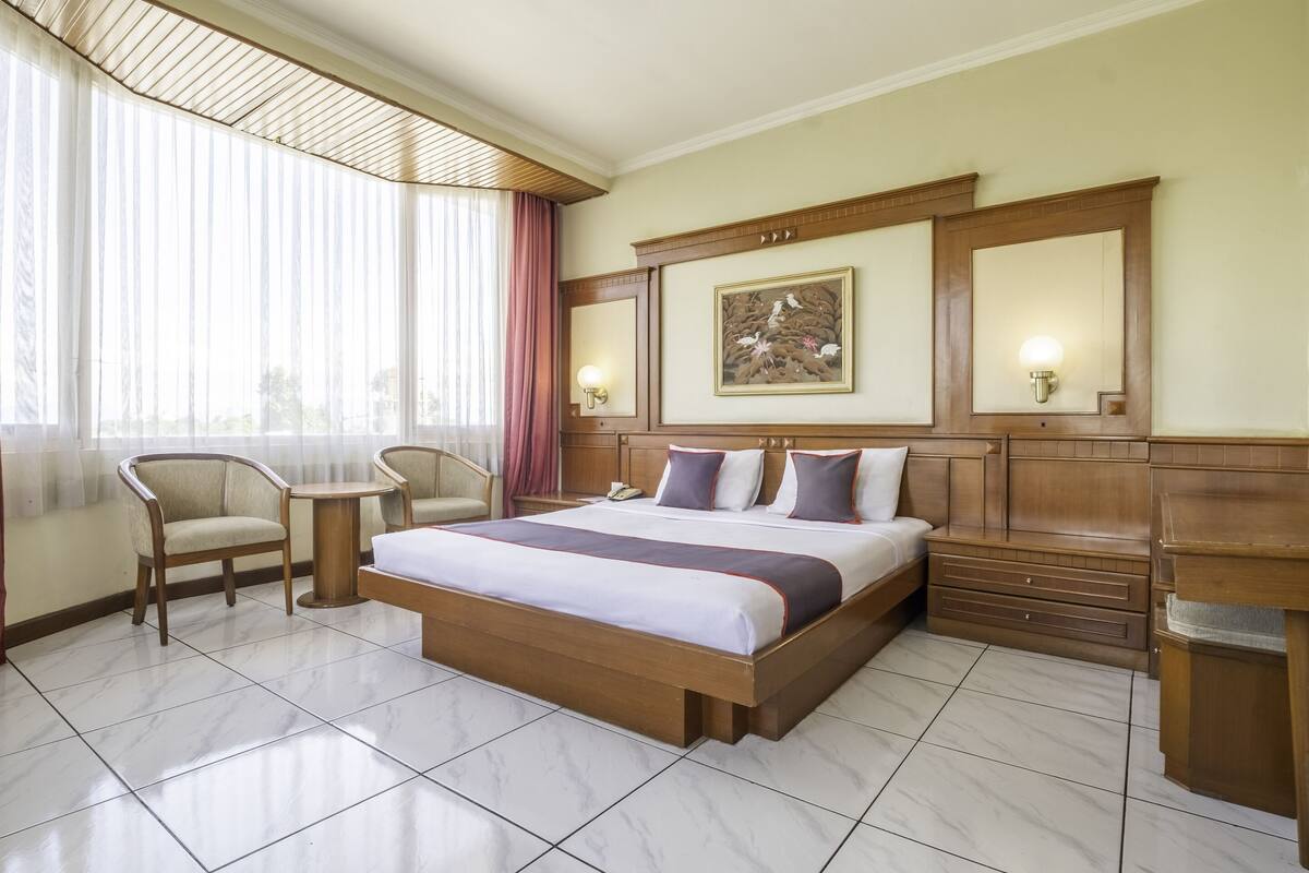 A hotel room with a large bed, a desk, two chairs, a small table, a nightstand, and a large window with curtains.