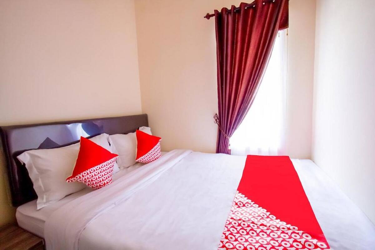 A neatly made bed with white bedding and red accents, a dark headboard, and a window with purple curtains.