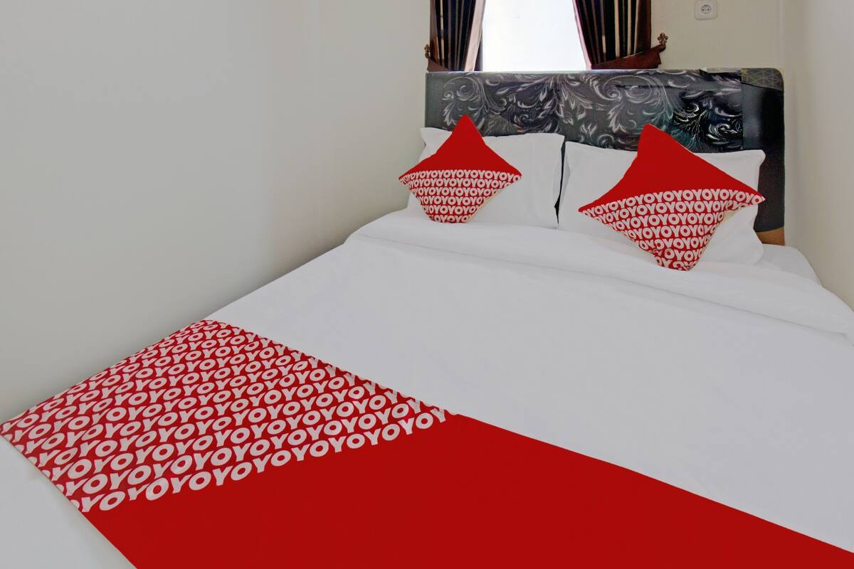 A bed with a white and red bedspread, two red pillows with a repeating pattern, and a headboard with a decorative design.