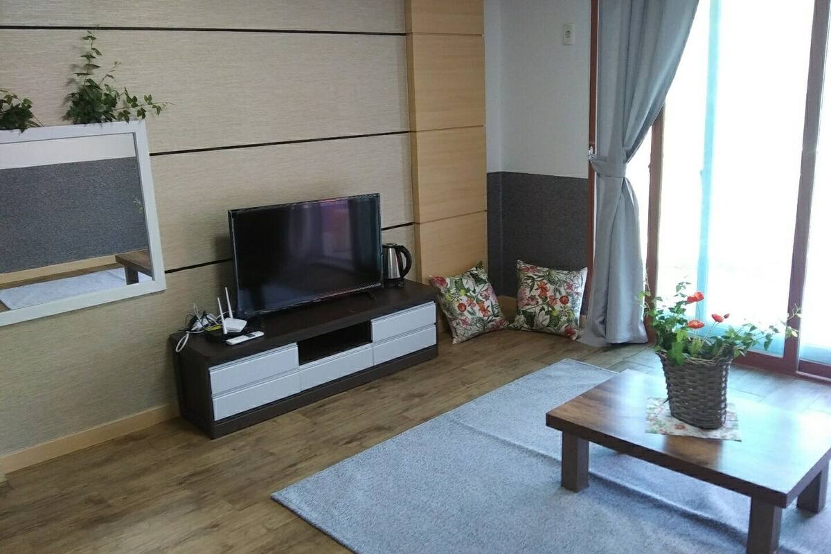 Een moderne woonkamer met een flatscreen televisie op een houten tv-meubel, een kleine tafel en een potplant.