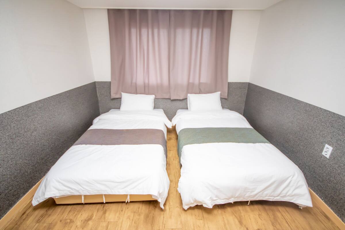 Een kamer met twee enkele bedden, een raam met dunne gordijnen en een houten vloer.