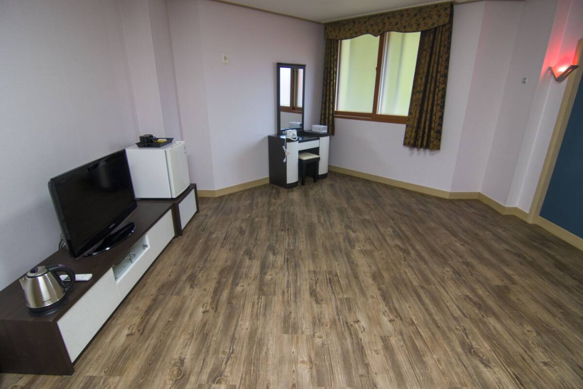 Een kamer met een flatscreen televisie op een houten tv-meubel, een klein wit bureau met een stoel, en een raam met gordijnen.