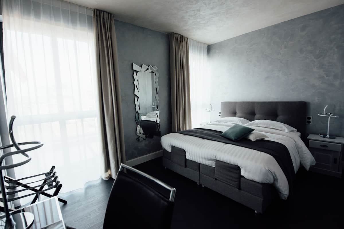 Quarto de hotel moderno com uma cama grande, mesa de cabeceira, uma cadeira e uma janela ampla com cortinas.