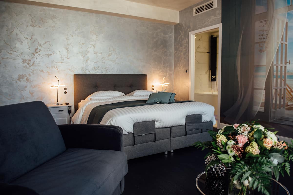 Quarto moderno com uma cama grande, mesas de cabeceira, um sofá e um vaso decorativo com flores.