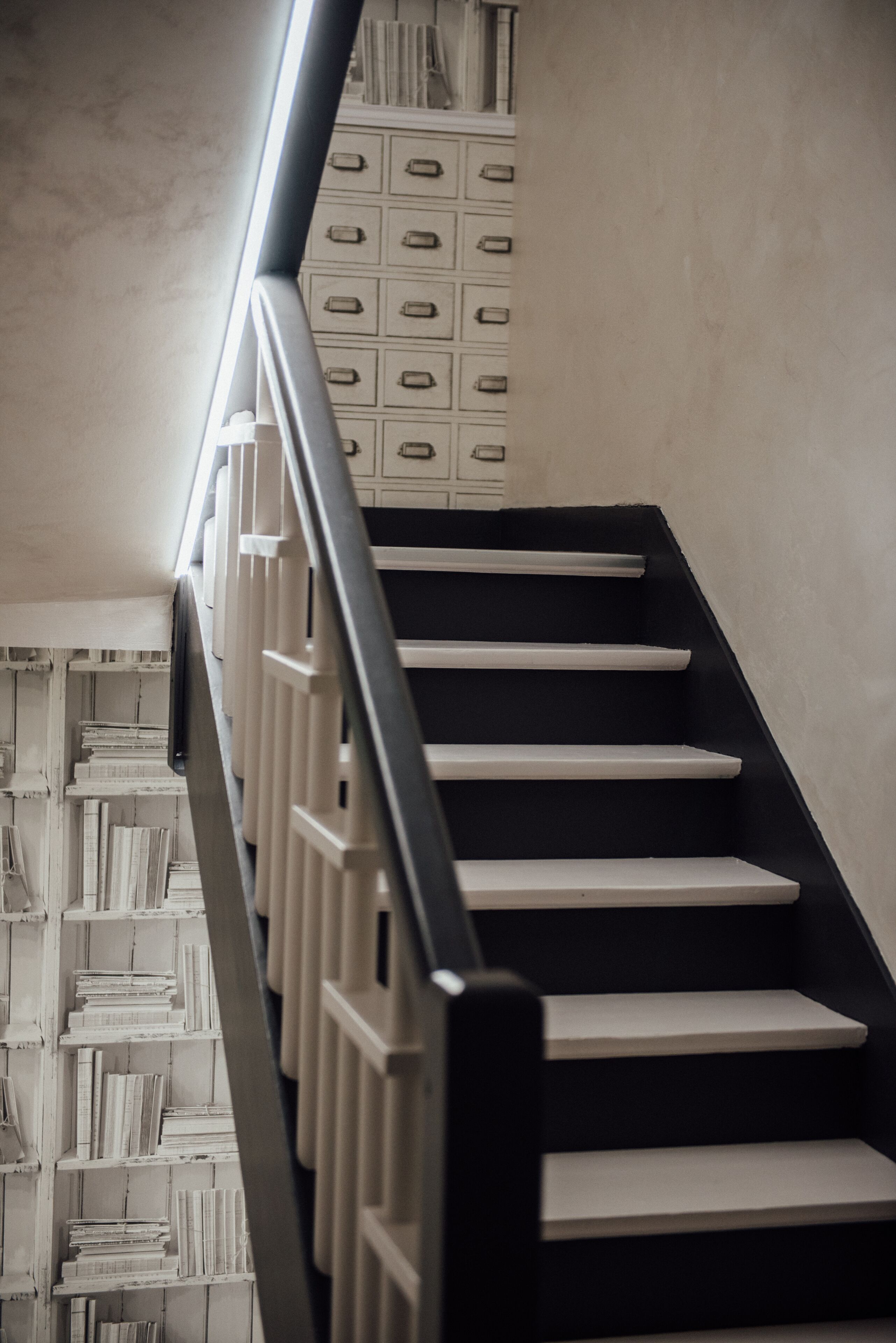 Een trap met een zwarte leuning en witte treden, leidend naar een kamer met een witte boekenkast.