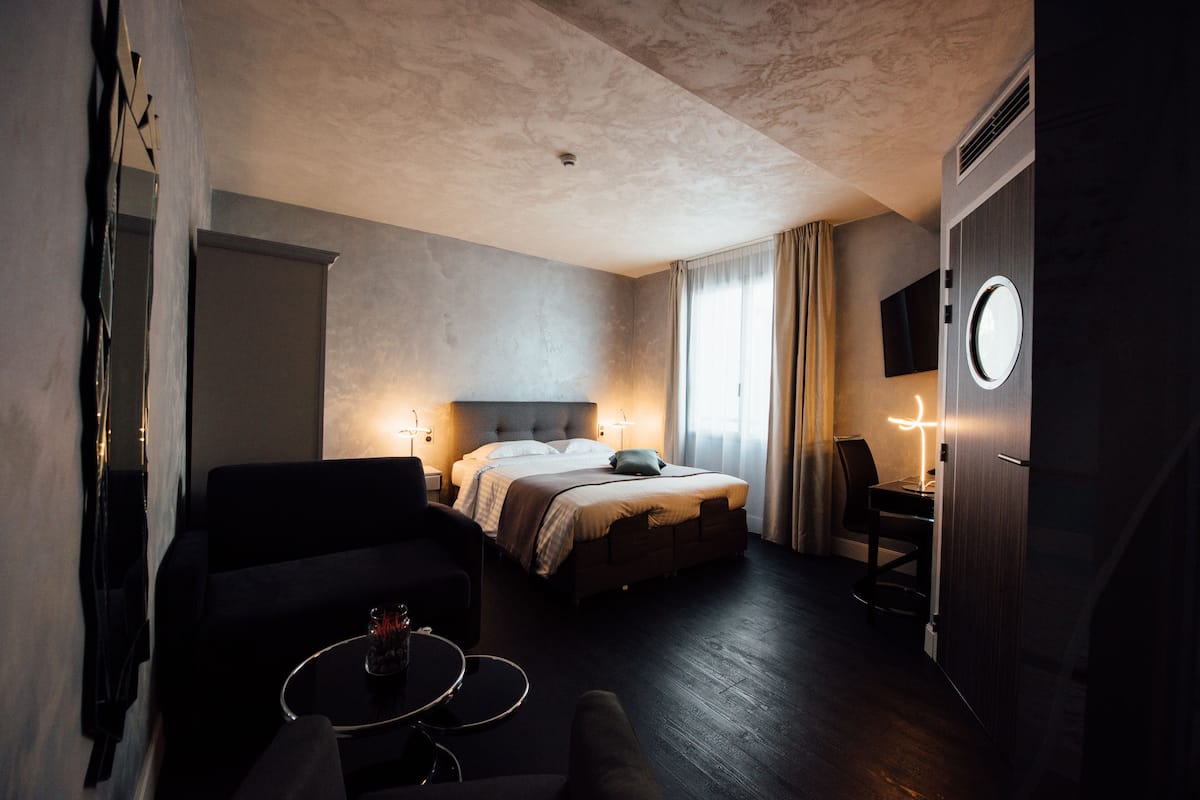 Quarto de hotel com cama, escrivaninha, cadeira, televisão e espelho redondo.