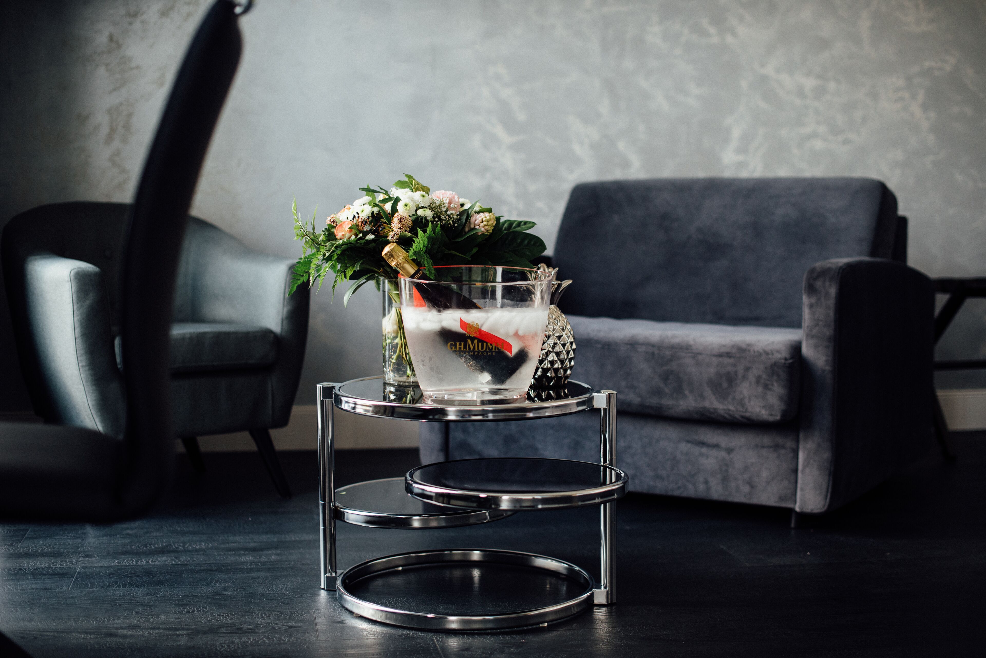 Uma sala de estar moderna com um sofá cinzento, uma poltrona de couro preto e uma mesa lateral metálica de três andares, com uma garrafa de champanhe e flores.