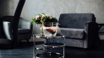 Uma sala de estar moderna com um sofá cinzento, uma poltrona de couro preto e uma mesa lateral metálica de três andares, com uma garrafa de champanhe e flores.