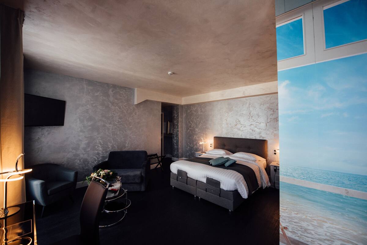 Quarto de hotel moderno com uma cama grande, uma escrivaninha e uma cadeira. Vê-se o oceano através de uma janela.