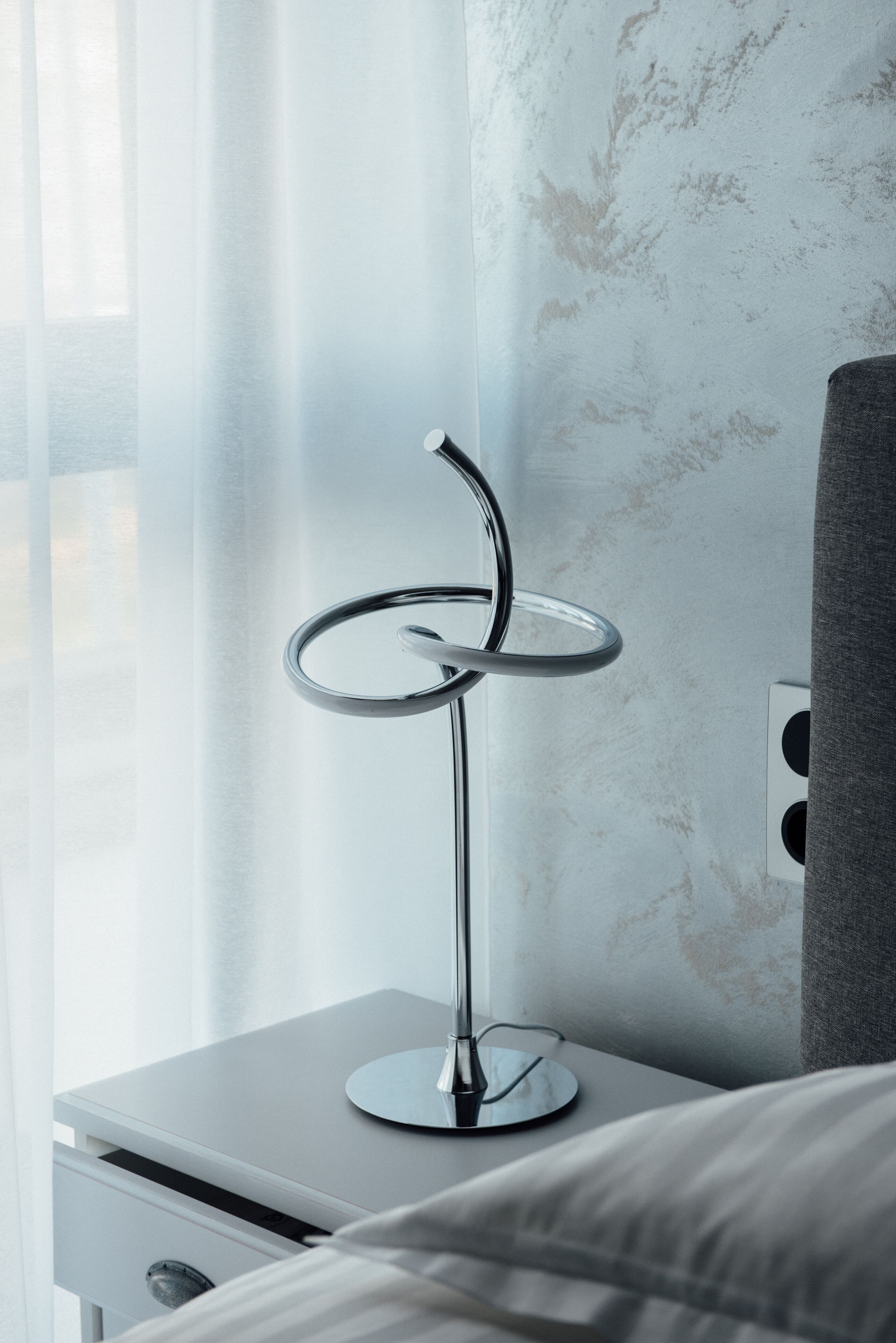A modern room with a bedside table, a lamp, and a window with sheer curtains.