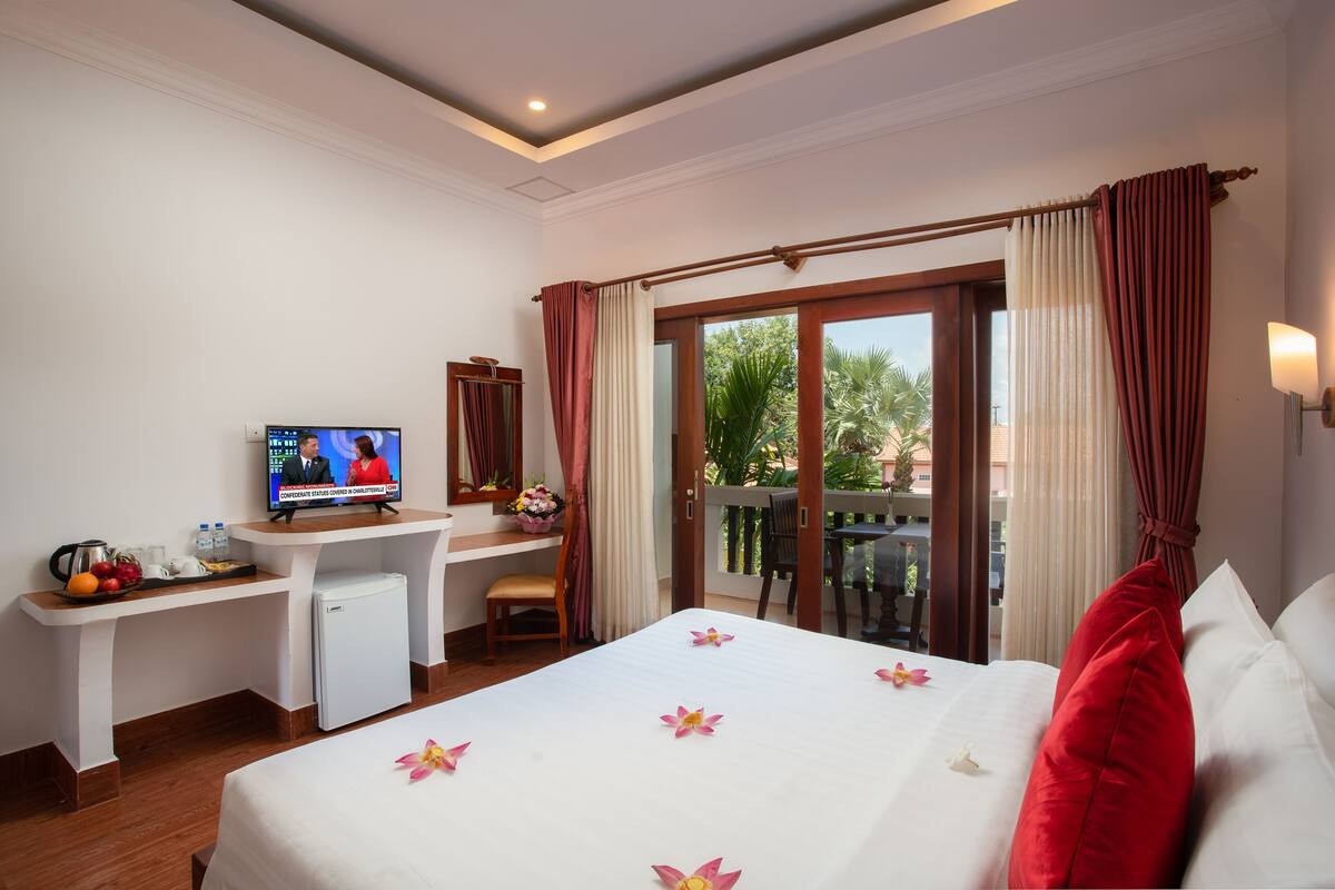 Habitación de hotel con cama, escritorio con televisor, refrigerador pequeño, silla y balcón con cortinas.