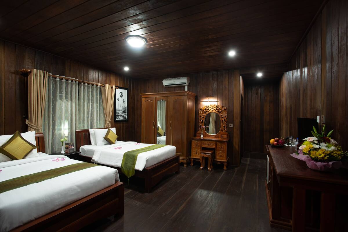 Habitación de hotel con dos camas, un armario de madera, un tocador con espejo y un escritorio con un jarrón de flores.