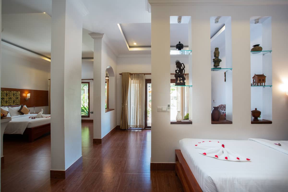 Habitación de hotel con piso de madera, una cama con ropa blanca, una mesita de noche, una lámpara y una ventana con cortinas.