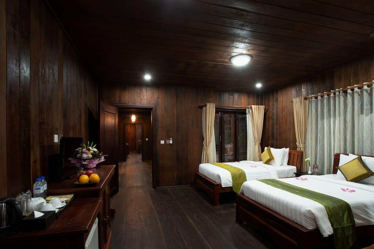 Habitación de hotel con paneles de madera, dos camas con ropa de cama verde y blanca, un escritorio con un jarrón de flores y una mesita de noche con una lámpara.