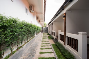 A narrow pathway flanked by lush greenery and a building with a tiled roof and white walls.