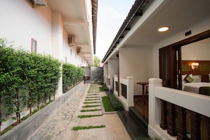 A narrow pathway flanked by lush greenery and a stone-tiled floor, leading to a building with a traditional wooden railing and a bed with a green pillow.