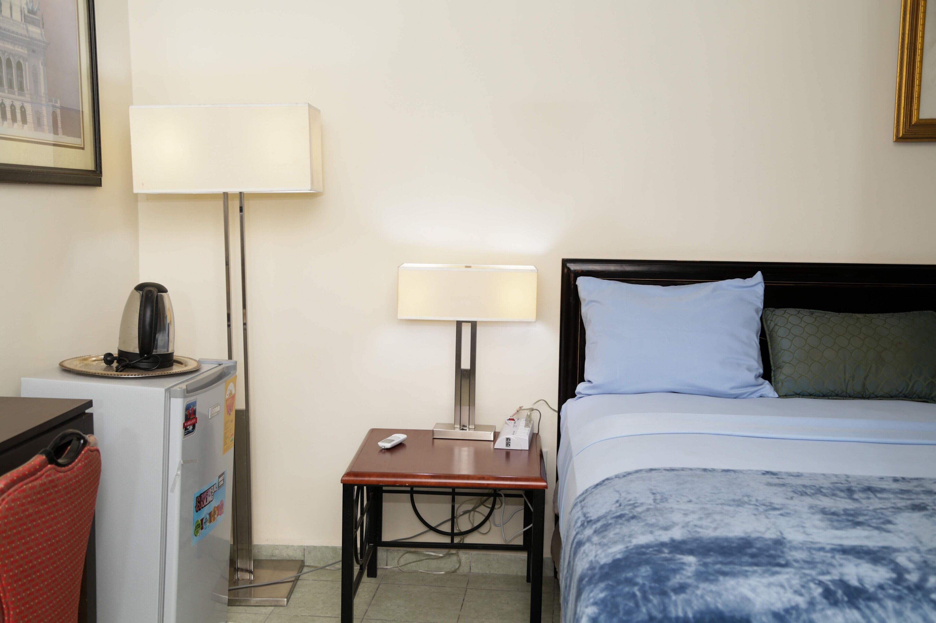 A hotel room with a bed, a desk with a lamp, a refrigerator, and a small table.