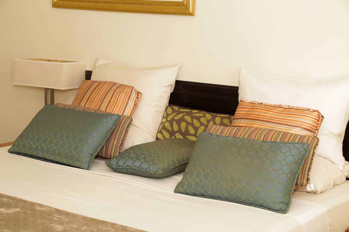 A neatly made bed with multiple pillows in various patterns and colors, a bedside lamp, and a framed picture above the bed.