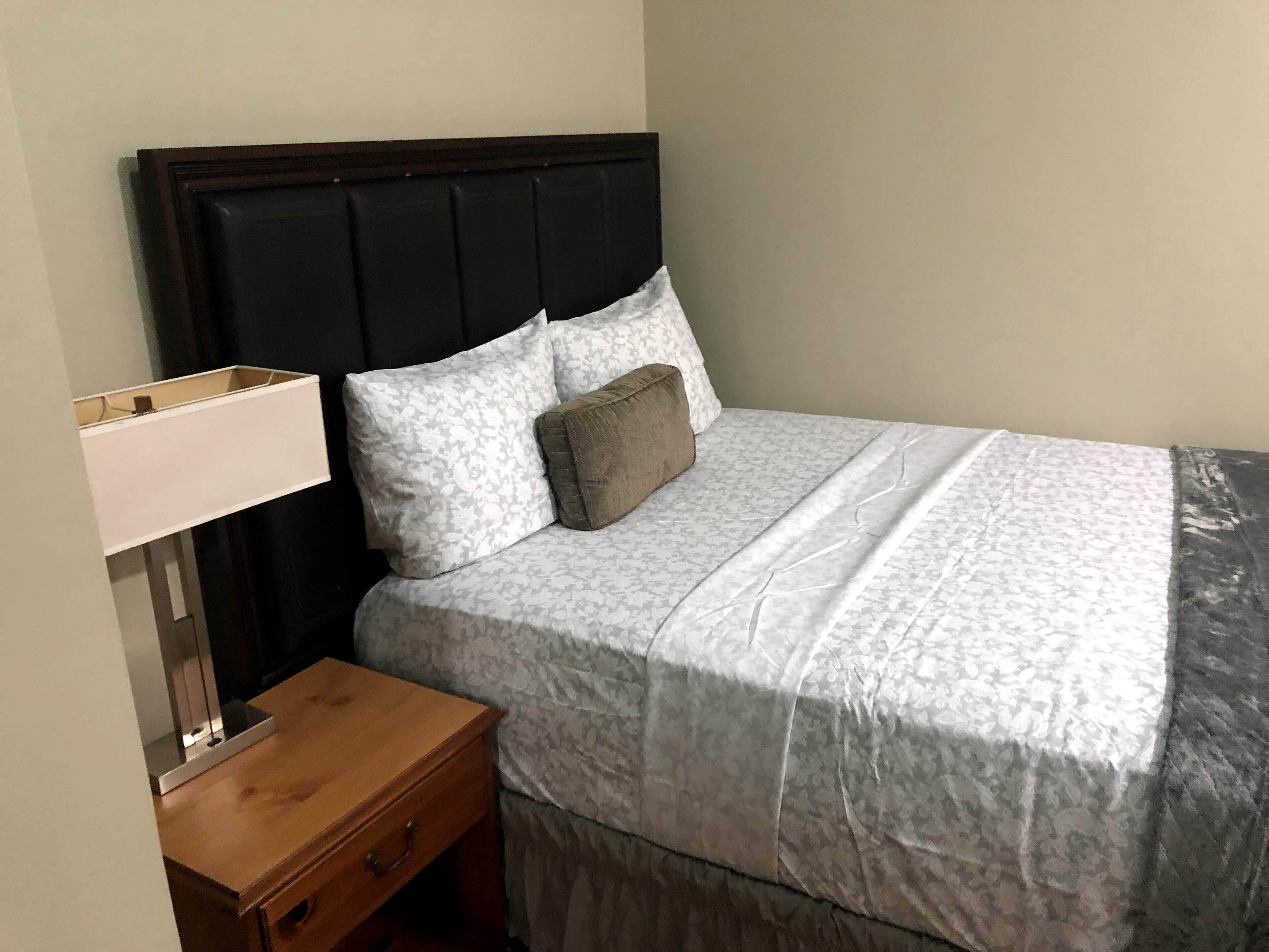 Una cama bien tendida con una colcha estampada y almohadas, una mesita de noche de madera con una lámpara, y un cabecero oscuro.