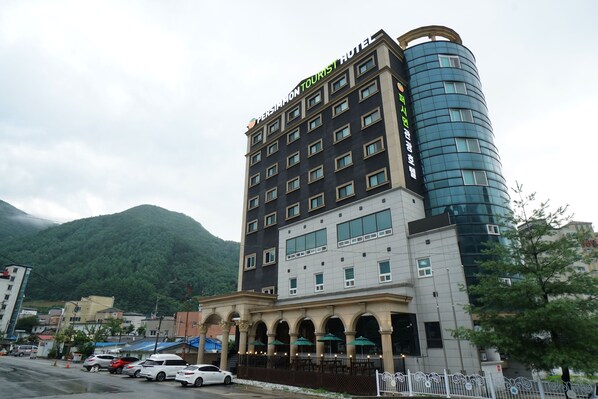 L’hôtel Persimmon Tourist, doté d’un design moderne, se distingue par sa façade aux couleurs contrastées, alternant le noir et le blanc, une tour en verre aux lignes courbes et une entrée située au niveau du sol, agrémentée d’un espace extérieur couvert avec des sièges.