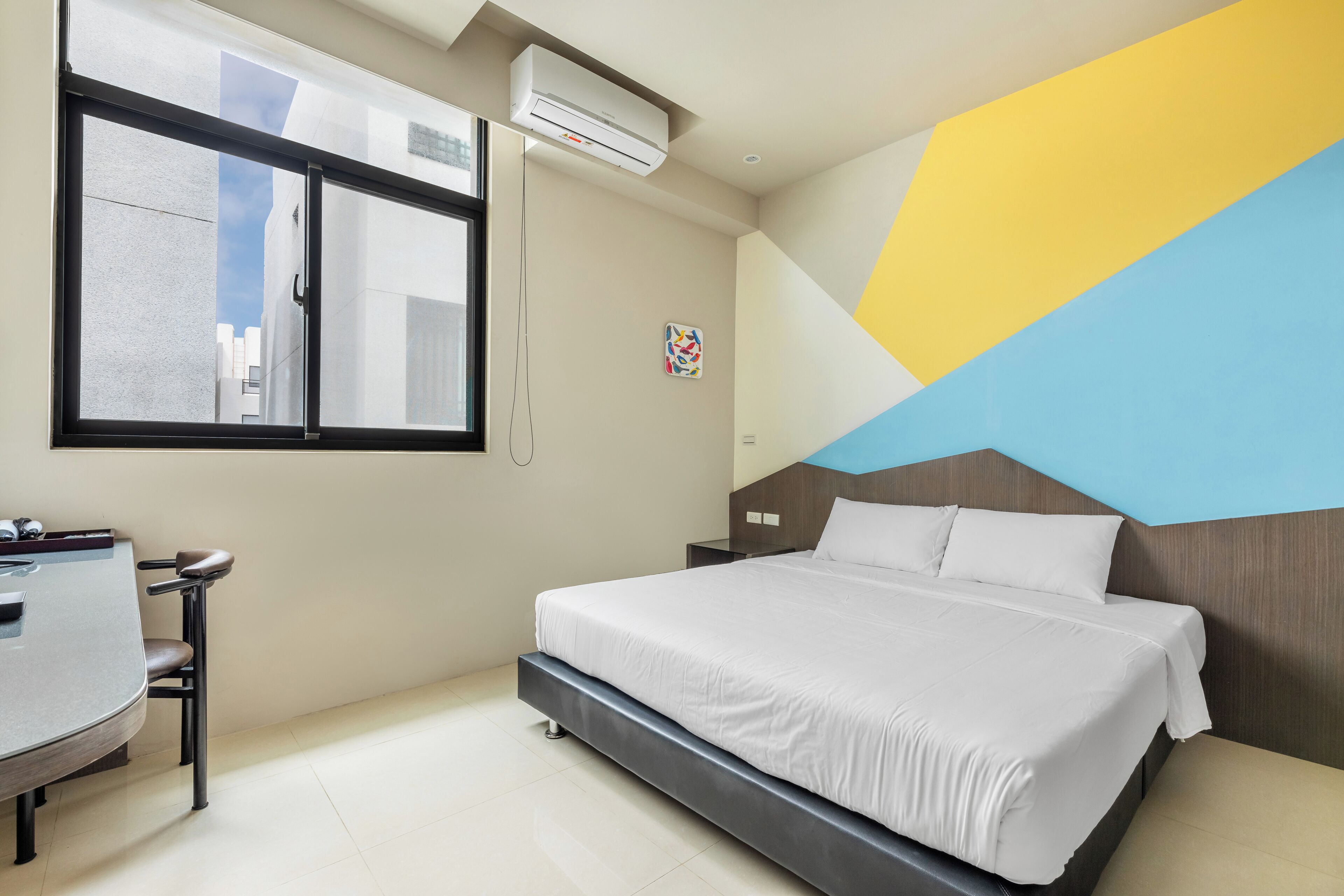 Ein modernes Hotelzimmer mit einem großen Bett, einem Schreibtisch mit Stuhl, einem Fenster und einer Klimaanlage.