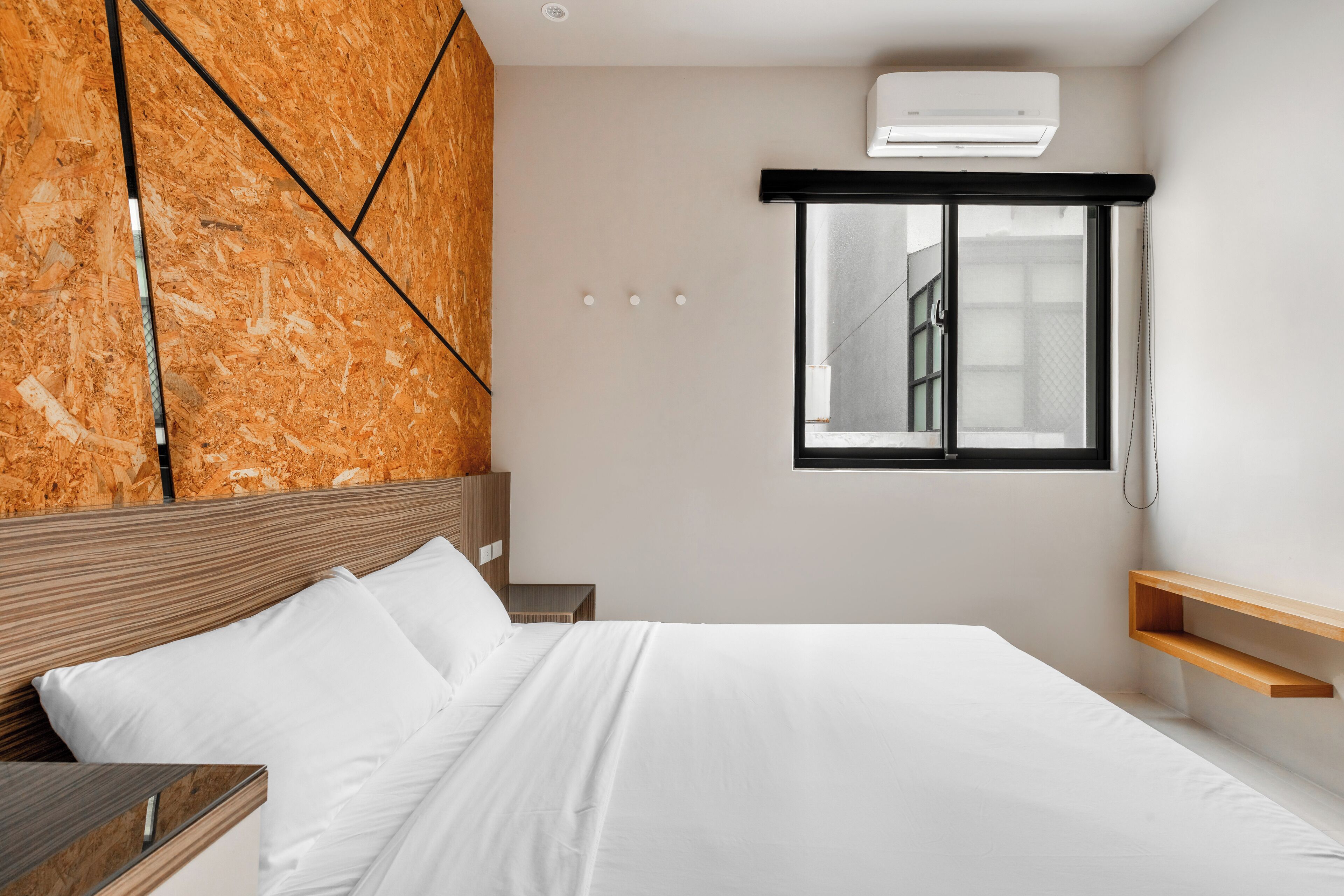 A modern bedroom with a wooden headboard, a bed with white linens, a window with a view, and a built-in air conditioning unit.