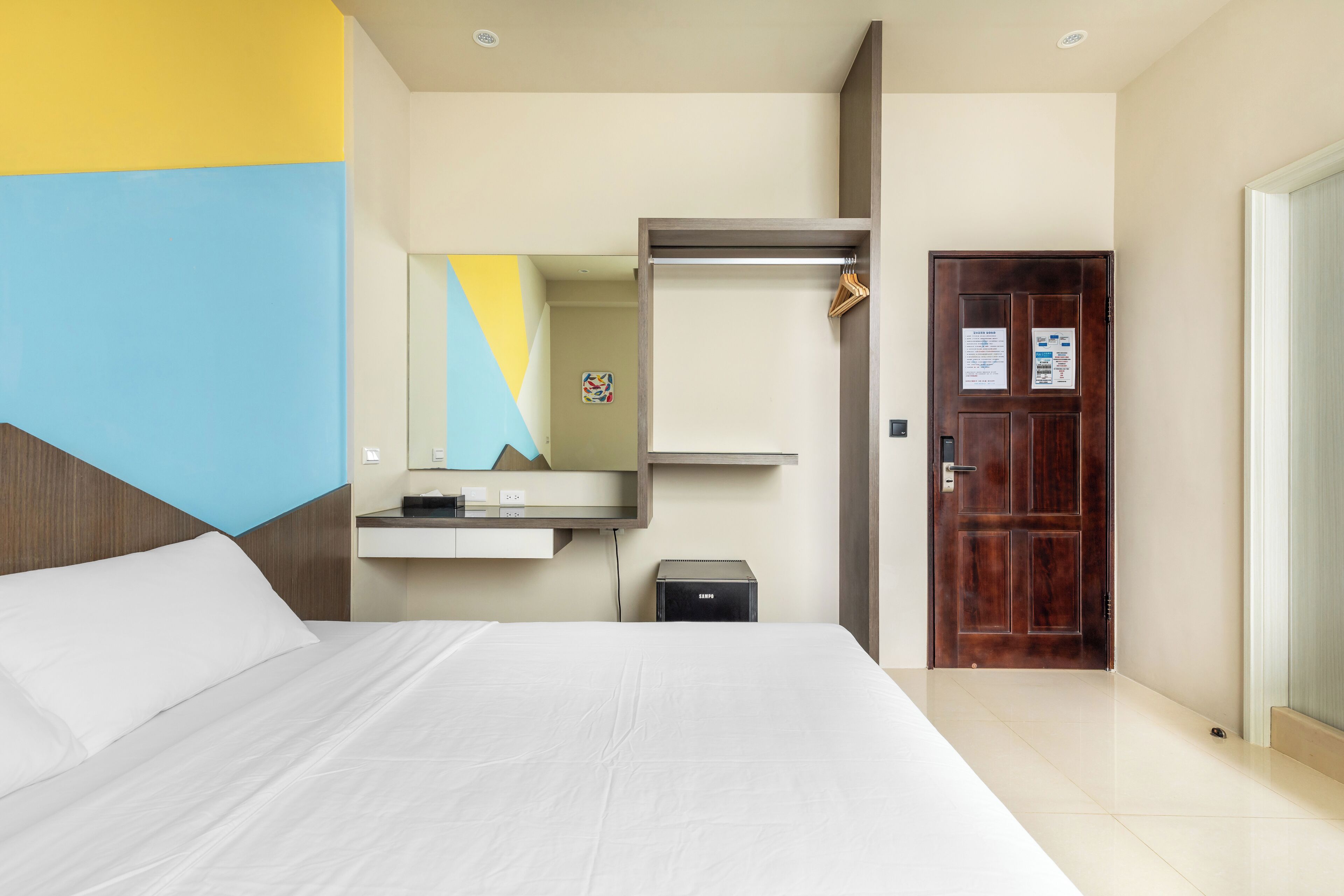 A hotel room with a large bed, a wardrobe, a small desk, and a door with a notice.