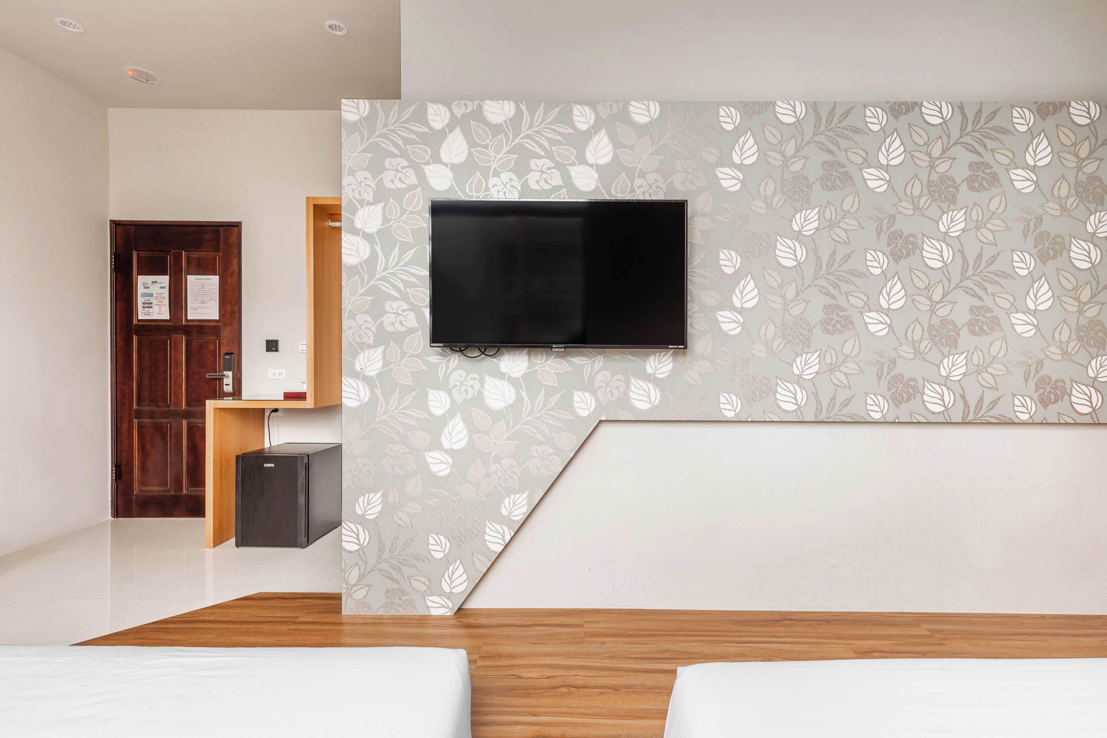 A hotel room with a flat-screen TV mounted on the wall, a wooden bed, a small desk with a computer, and a door with a bulletin board.